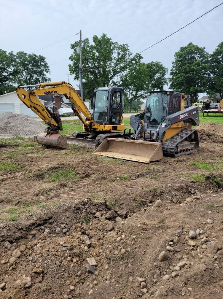 Two construction machines on a dirt lot, with an excavator and a skid steer near piles of soil.