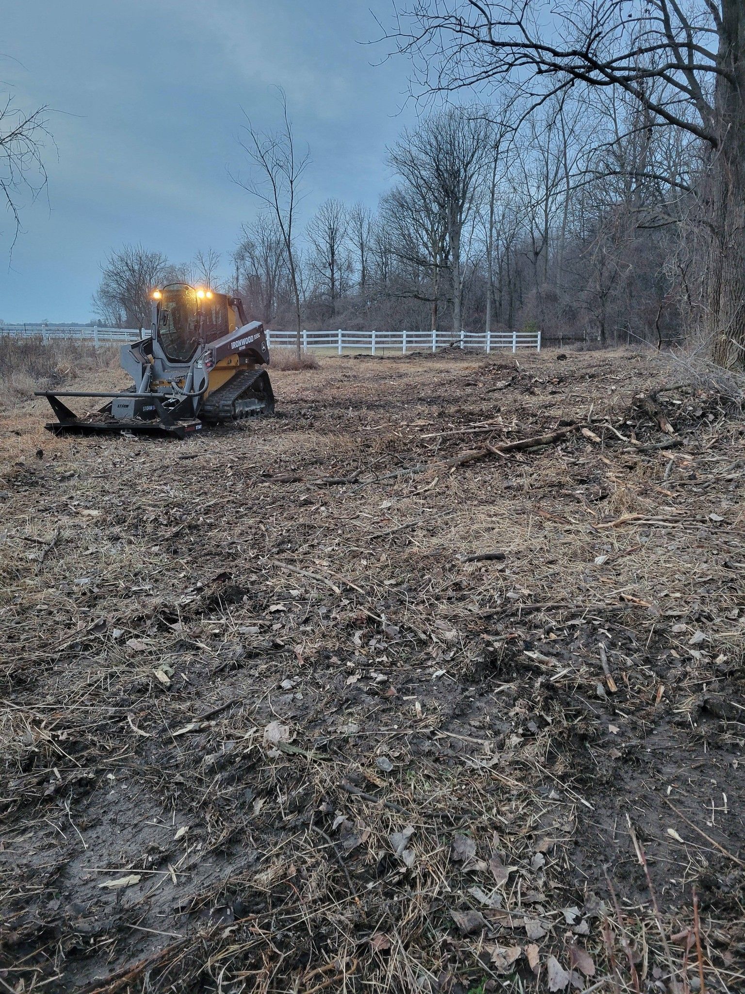 A small excavator clearing leaves in a wooded yard near a white fence at dusk.