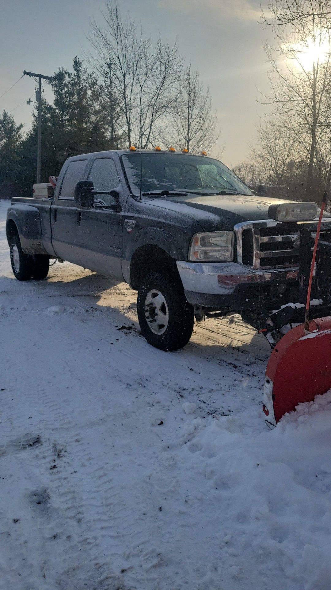 Gray pickup truck with plow parked on snowy ground at sunset, trees in the background