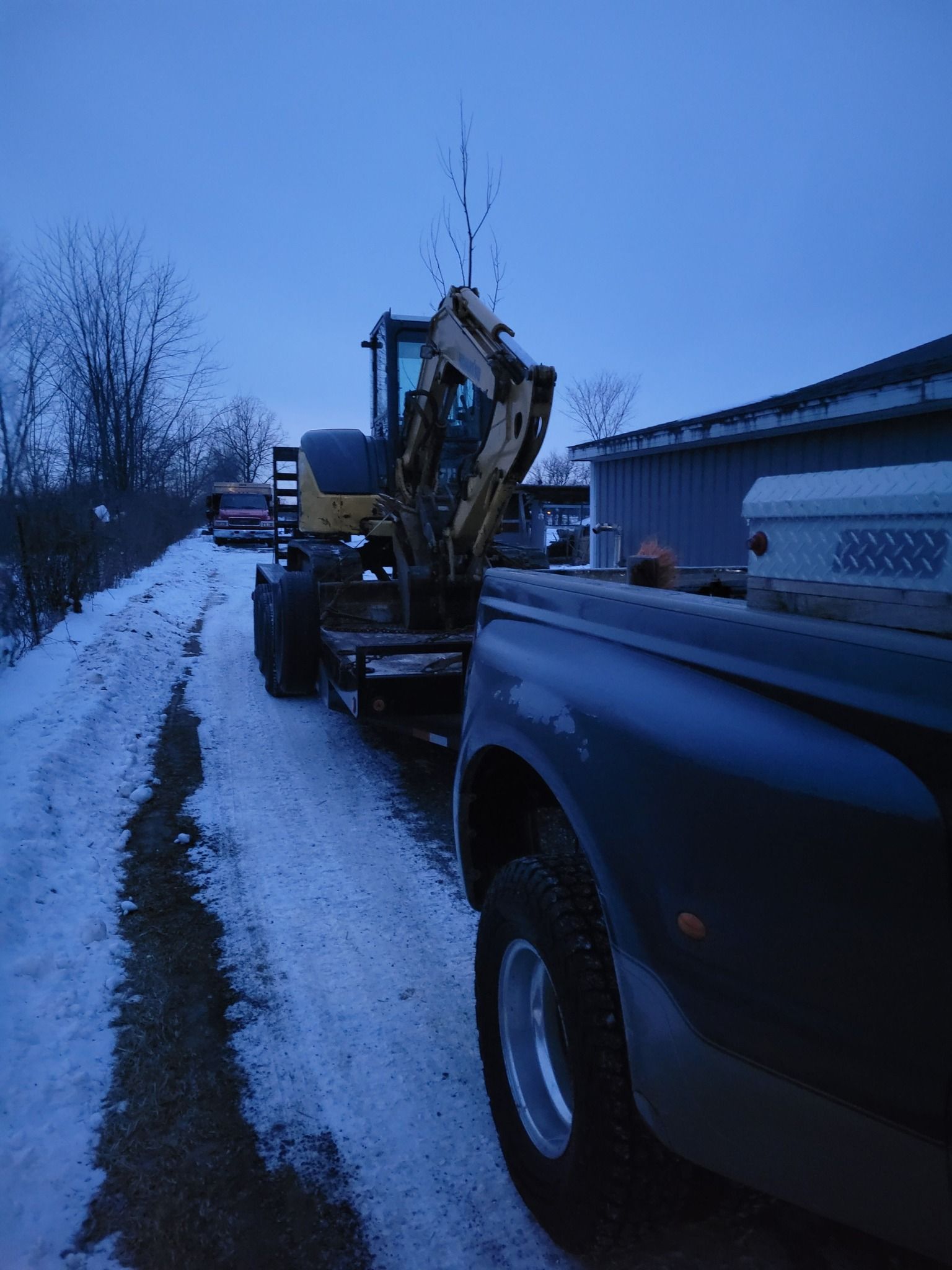 Pickup truck hauling a yellow excavator on a snowy roadside at dusk