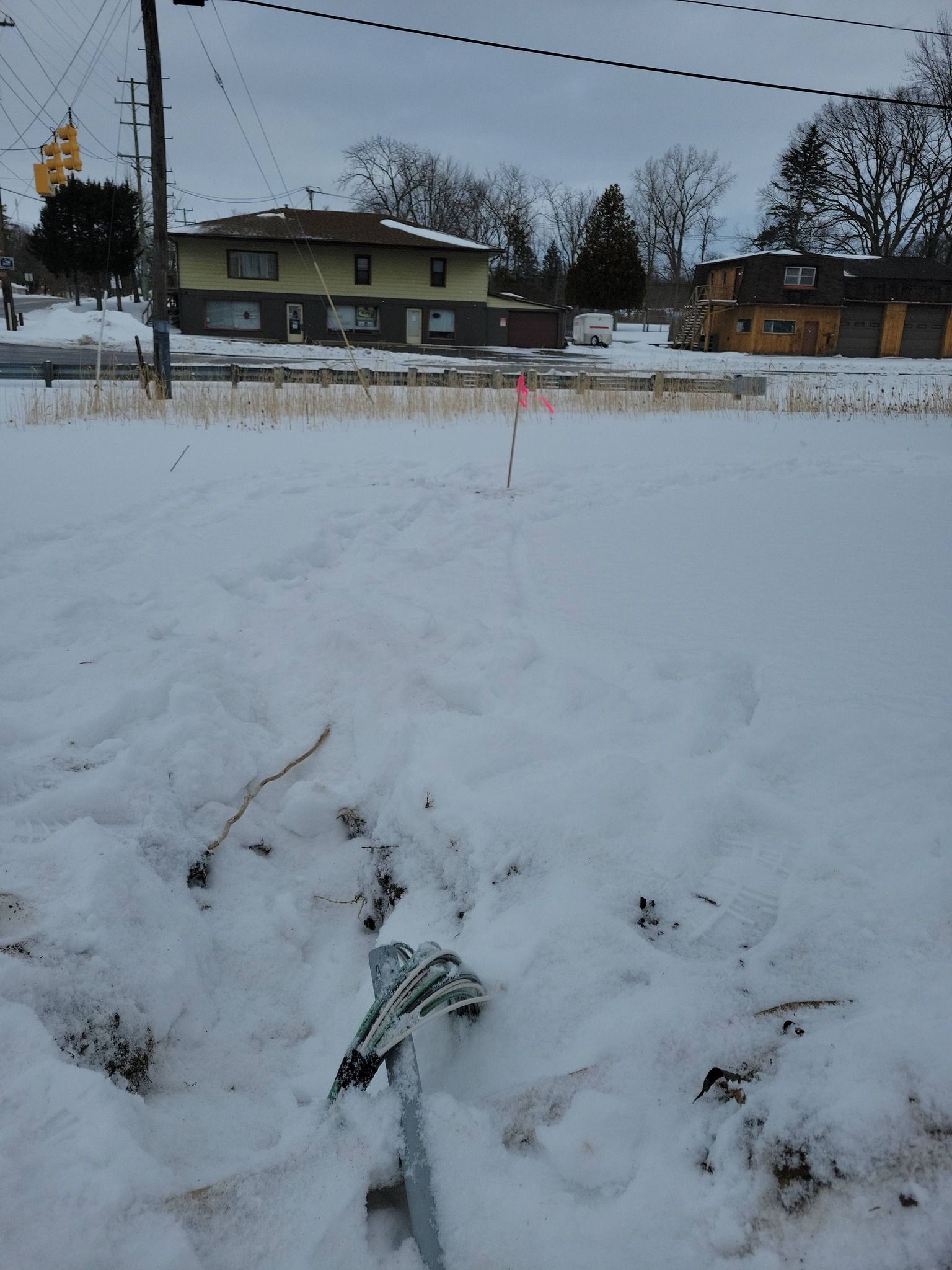 Snow-covered field with a red marker stake, houses in the background, and overcast winter sky.