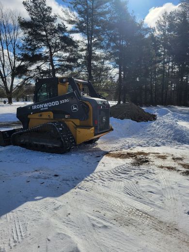 Yellow skid-steer loader clearing snow on a sunlit driveway beside trees