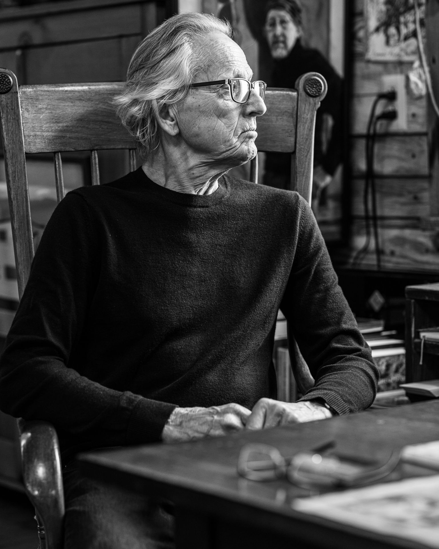 Black-and-white portrait of a seated person in glasses, looking left in a wooden chair indoors.