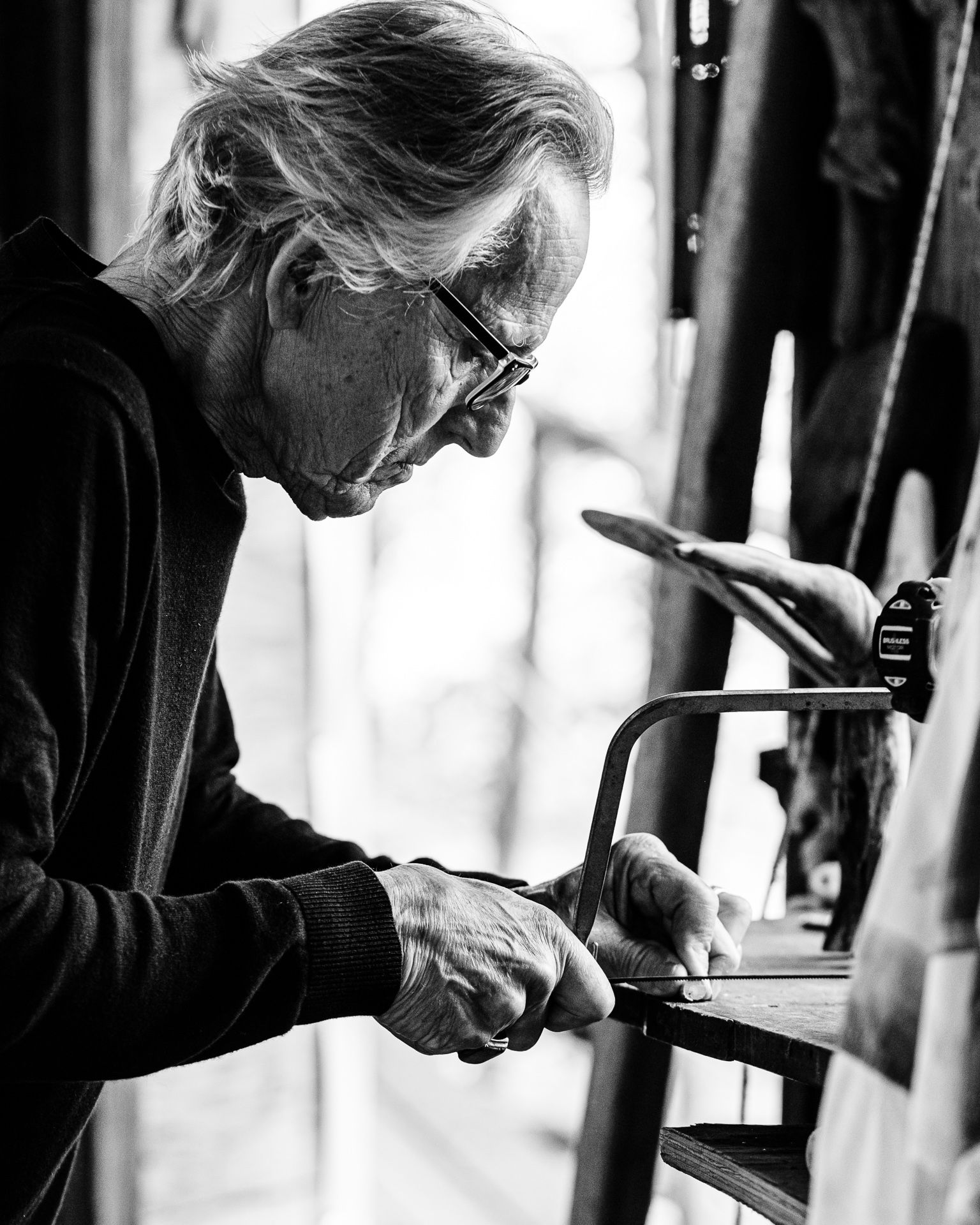 Elderly person using a hand saw at a workbench in a workshop, in black and white.