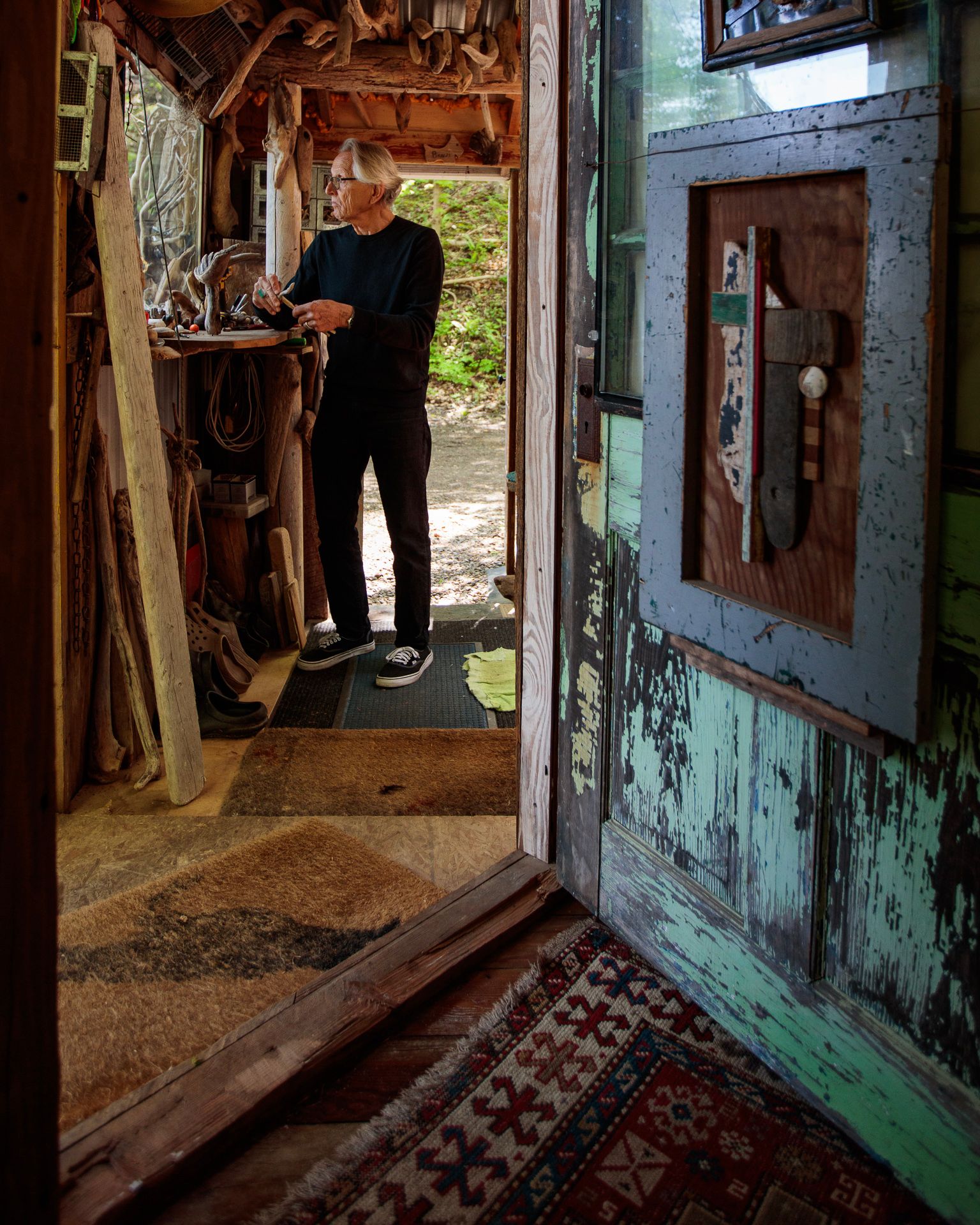 Person standing in a rustic doorway, holding an object, with weathered blue-green door and patterned rug nearby
