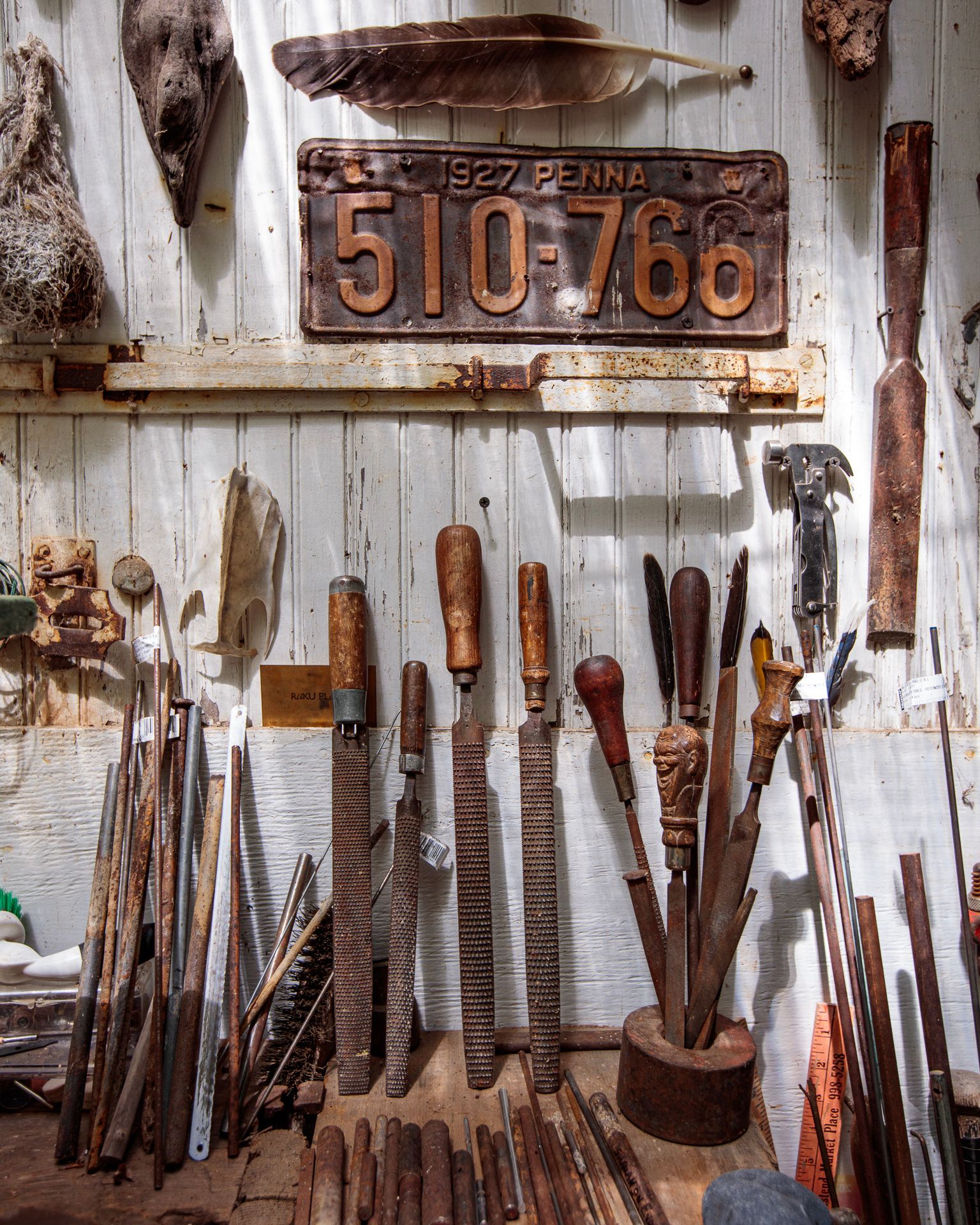 Rusty vintage tools and knives displayed on a weathered wall beneath an old metal sign.