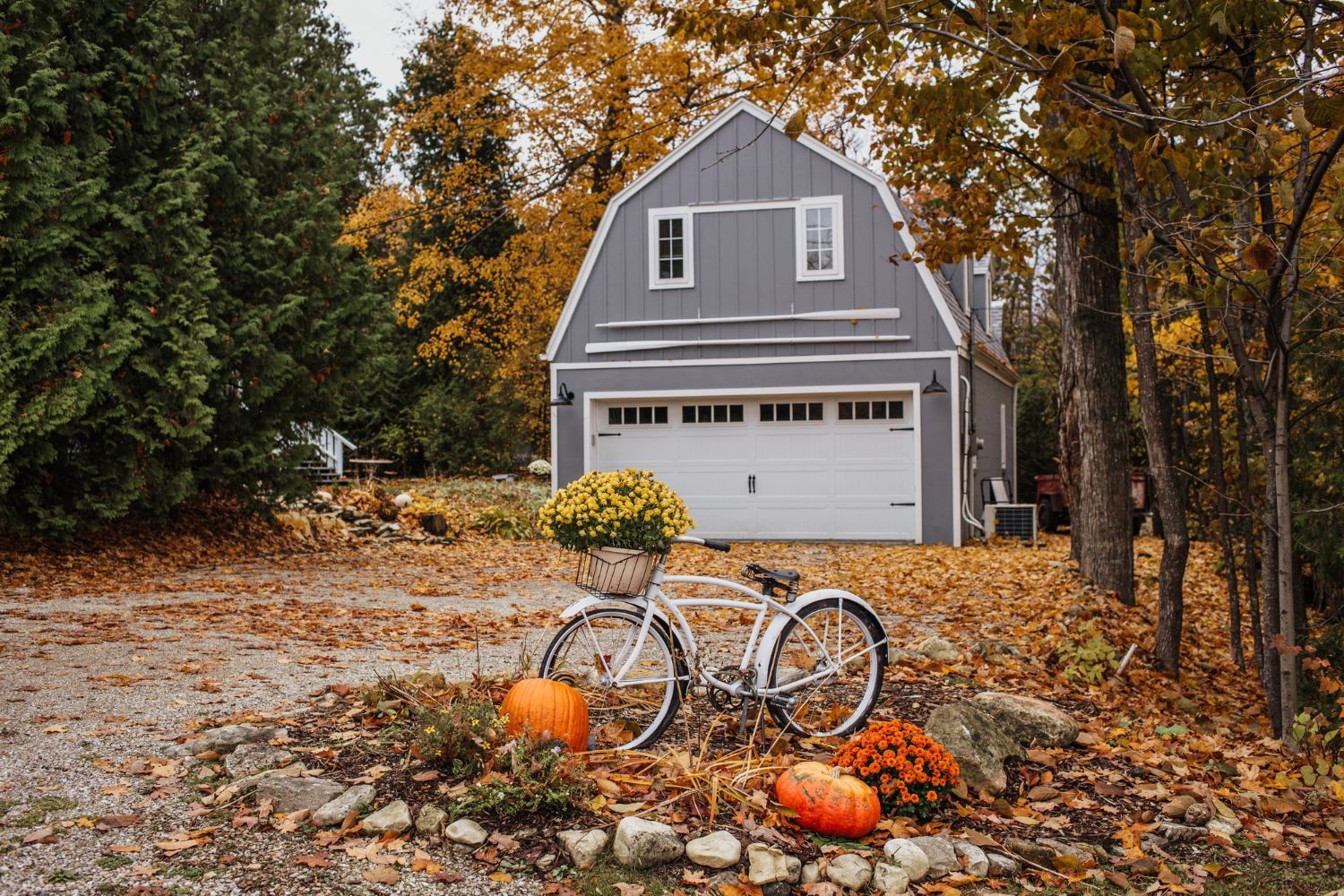 Gray house with garage amid autumn trees, with a bicycle and pumpkins in a leaf-covered yard.