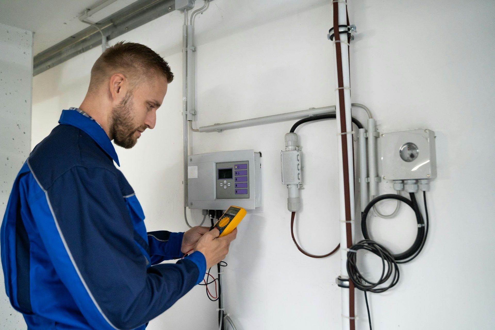 Technician using a handheld meter to inspect wall-mounted electrical equipment in a utility room