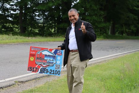 Man standing by roadside holding a colorful sign and giving a thumbs-up on a grassy verge