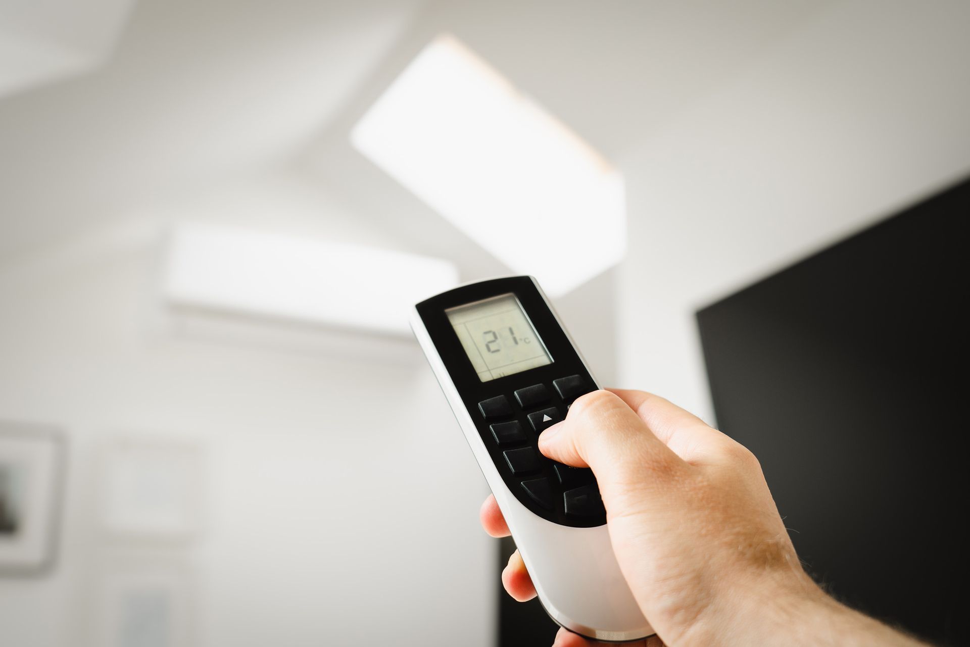 Hand holding a remote toward a ceiling with bright skylights and a wall-mounted TV
