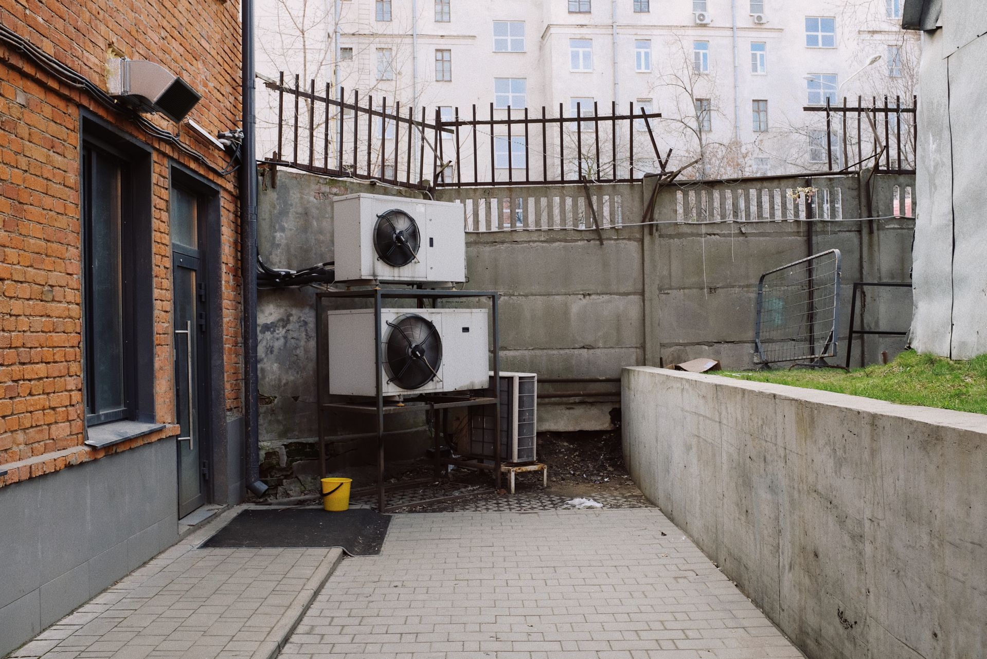 Narrow courtyard with two air-conditioning units, brick wall, concrete fence, and paved walkway.