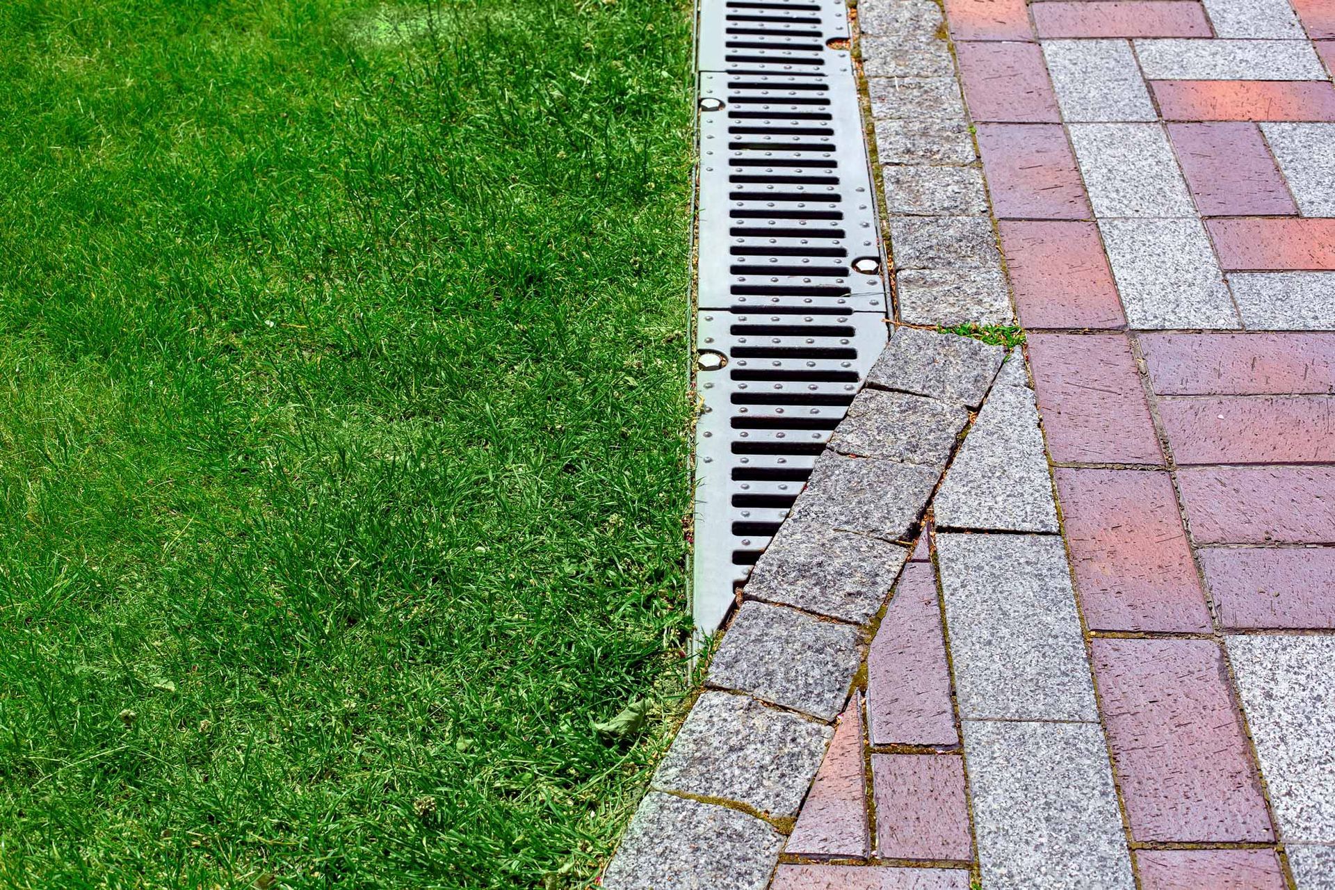 A drain on the side of a brick walkway next to a lush green lawn.