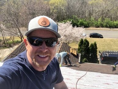Smiling man in sunglasses and cap on a rooftop, with workers and trees in the background.