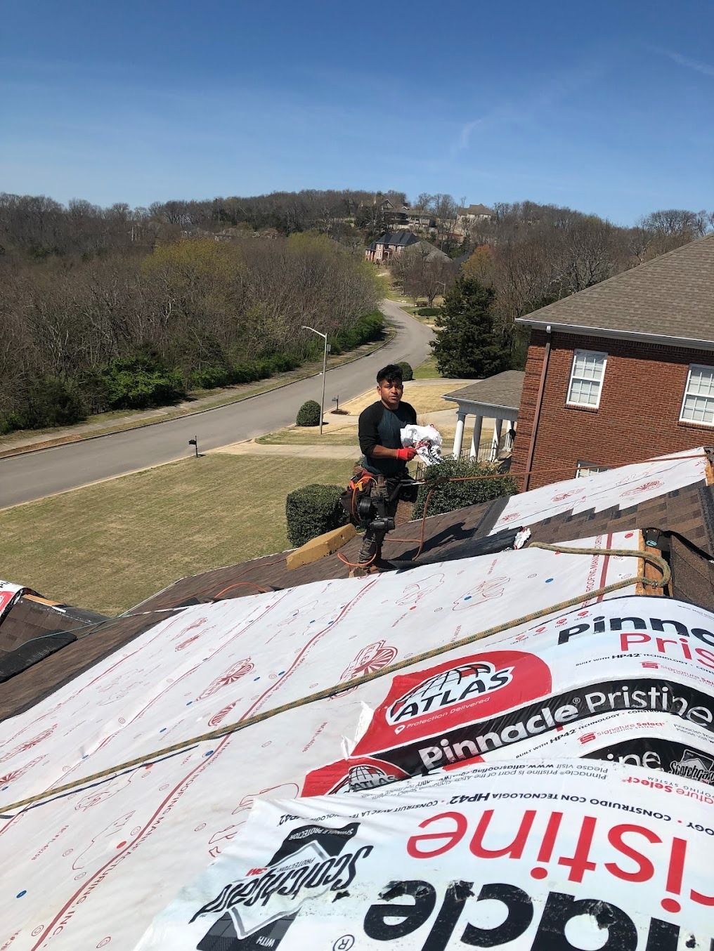 Worker on a steep roof installing underlayment beside a brick house on a clear day.