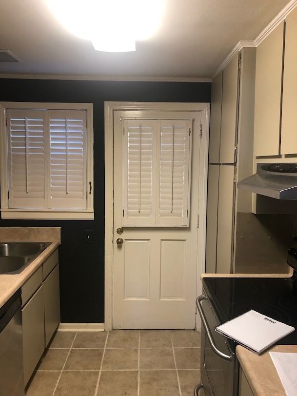 Narrow kitchen with beige cabinets, dark walls, tiled floor, and a back door with shutters.