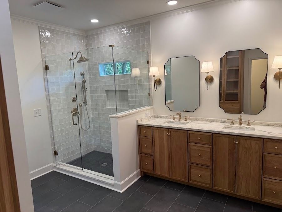 Spacious bathroom with glass shower, gray tile, and double vanity with wood cabinets and mirrors