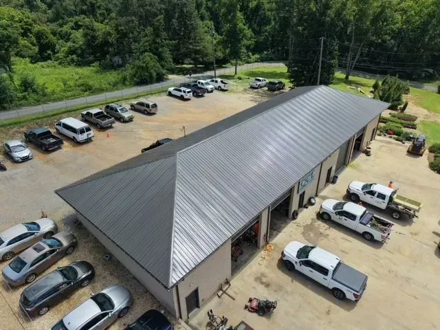 Aerial view of a large metal-roofed building with parked cars and trucks in a gravel lot beside trees.