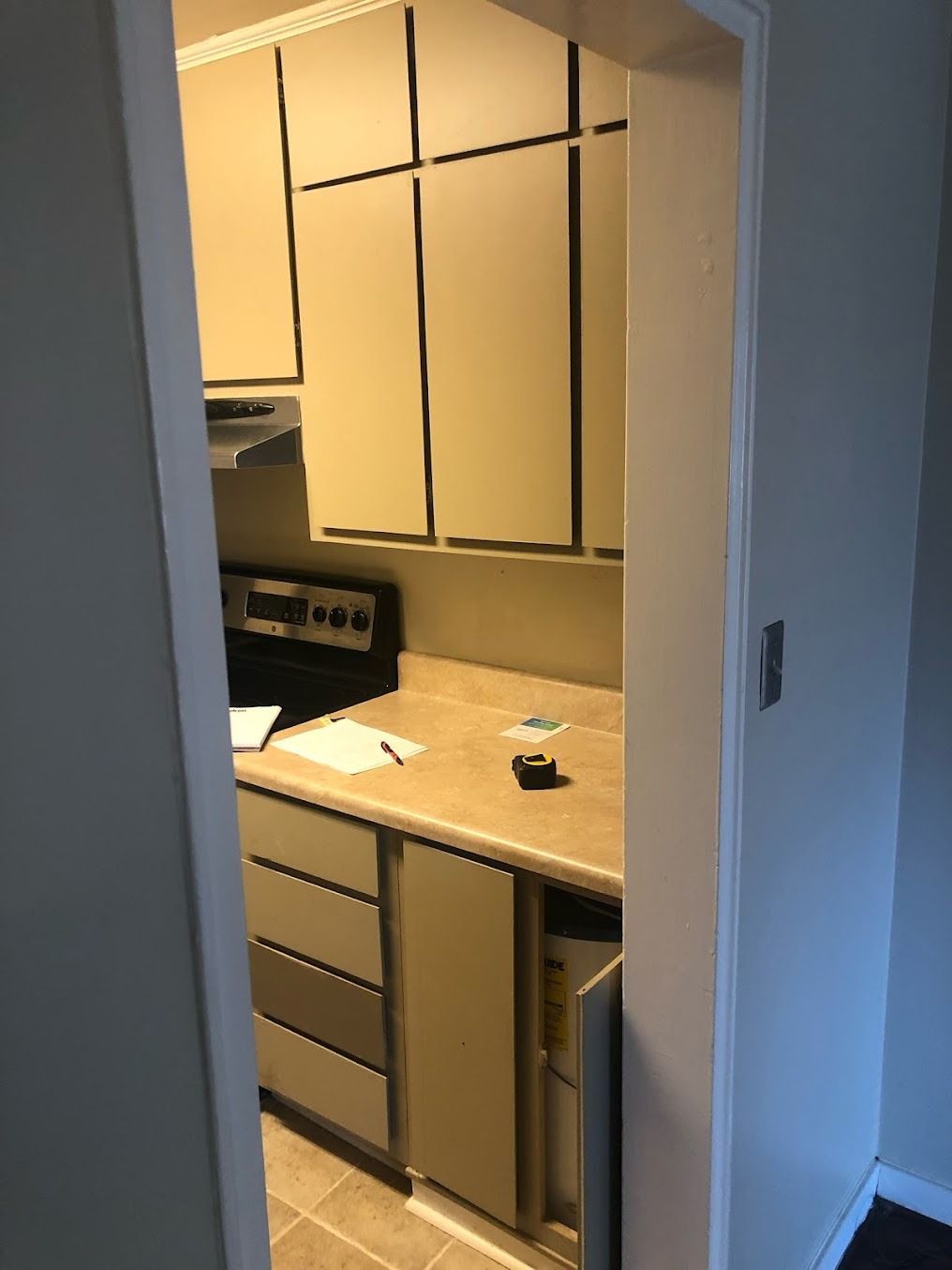 A small kitchenette with beige cabinets, countertop, drawers, and a doorway view into the room.