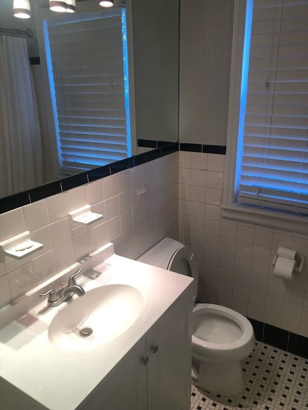 Small bathroom with sink, mirror, toilet, white tile, and windows with blinds.