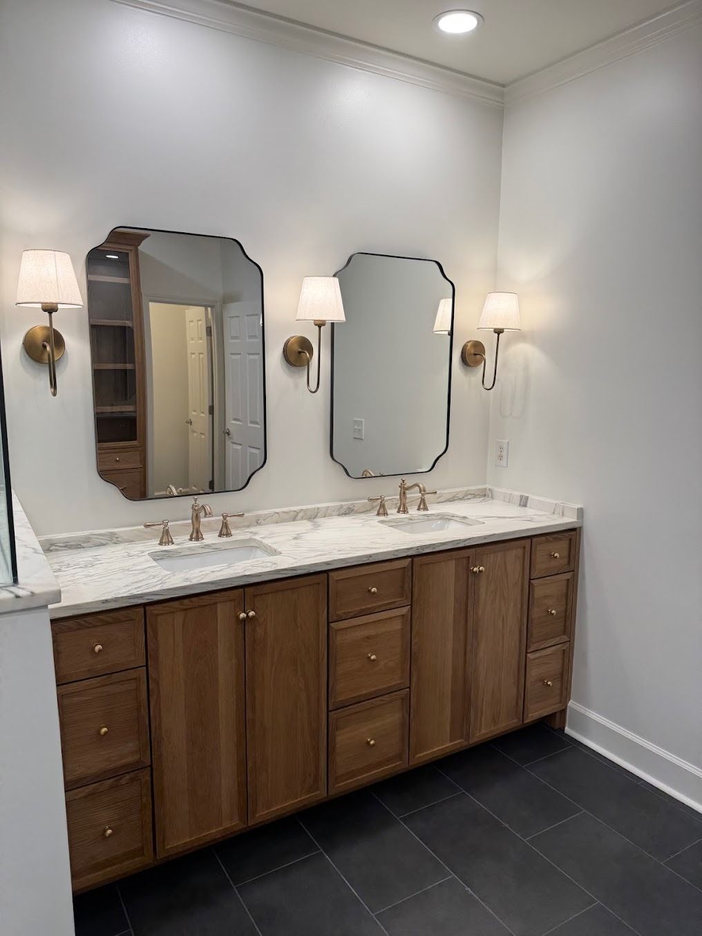 Bathroom vanity with two mirrors, wall sconces, marble countertop, and dark tile floor.