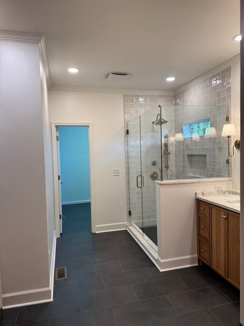 Bright bathroom with glass shower, white walls, dark tile floor, and wood vanity beside a blue doorway