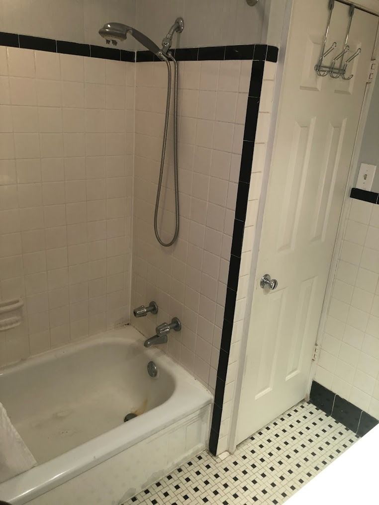 Small bathroom with a white tub-shower, black trim, and a white door beside a tiled floor.