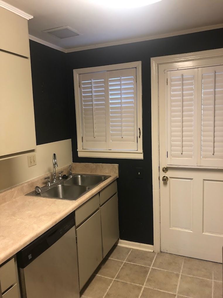 Small kitchen with beige counters, stainless sink, dark walls, white shutters, and a white door.