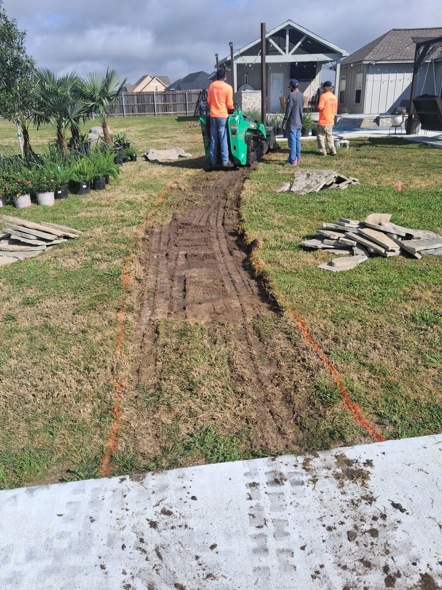 Workers laying sod in a muddy backyard with houses and construction materials nearby