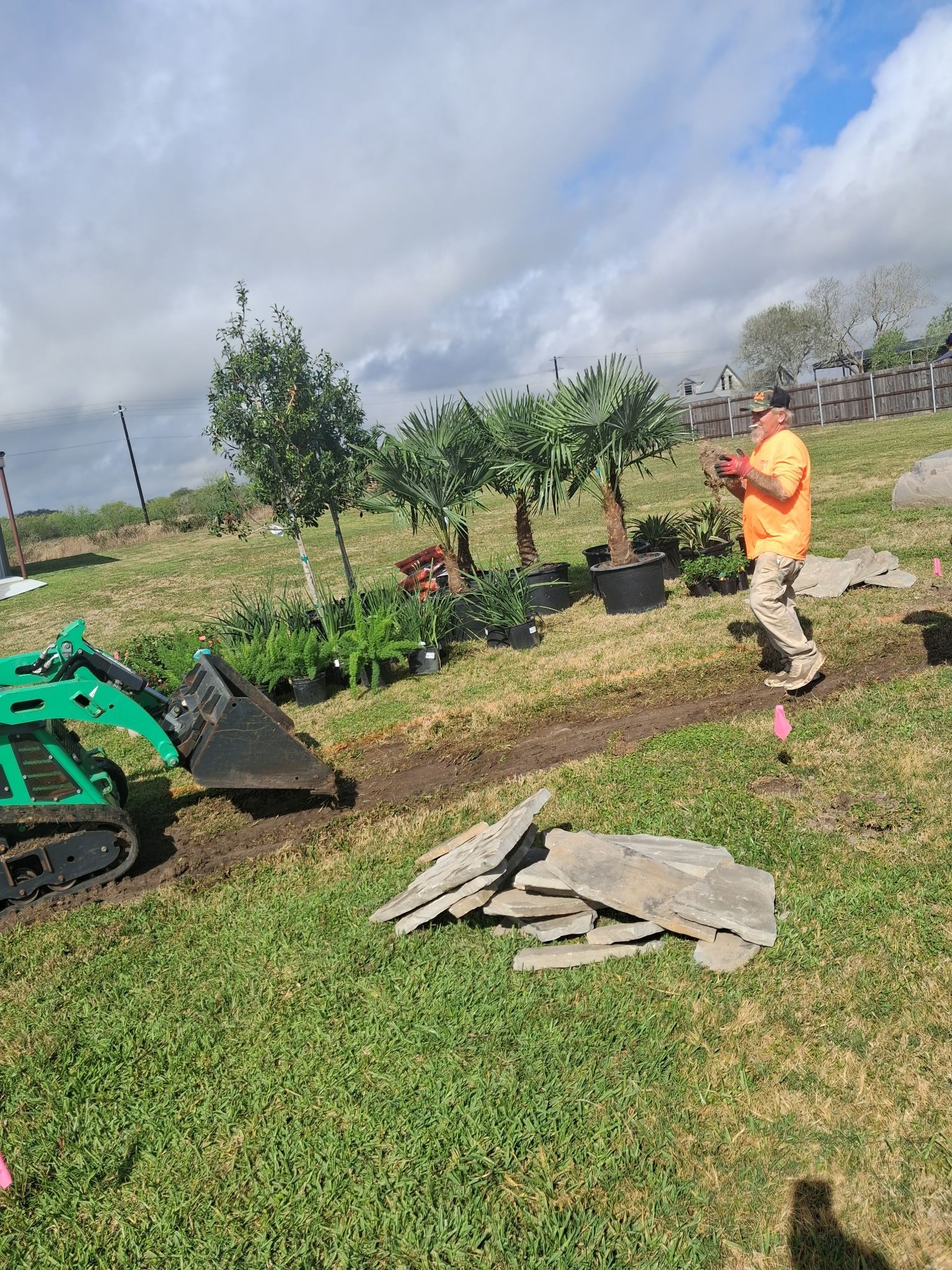 Person in orange works in a landscaped garden beside a small excavator under a cloudy sky.
