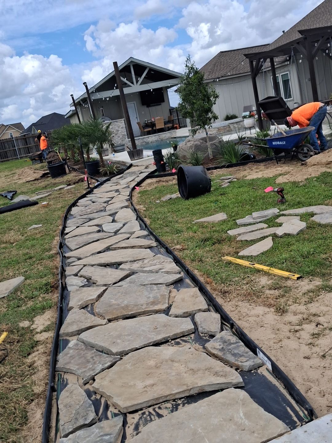 Stone walkway under construction leading to houses, with workers and scattered materials on grassy ground.