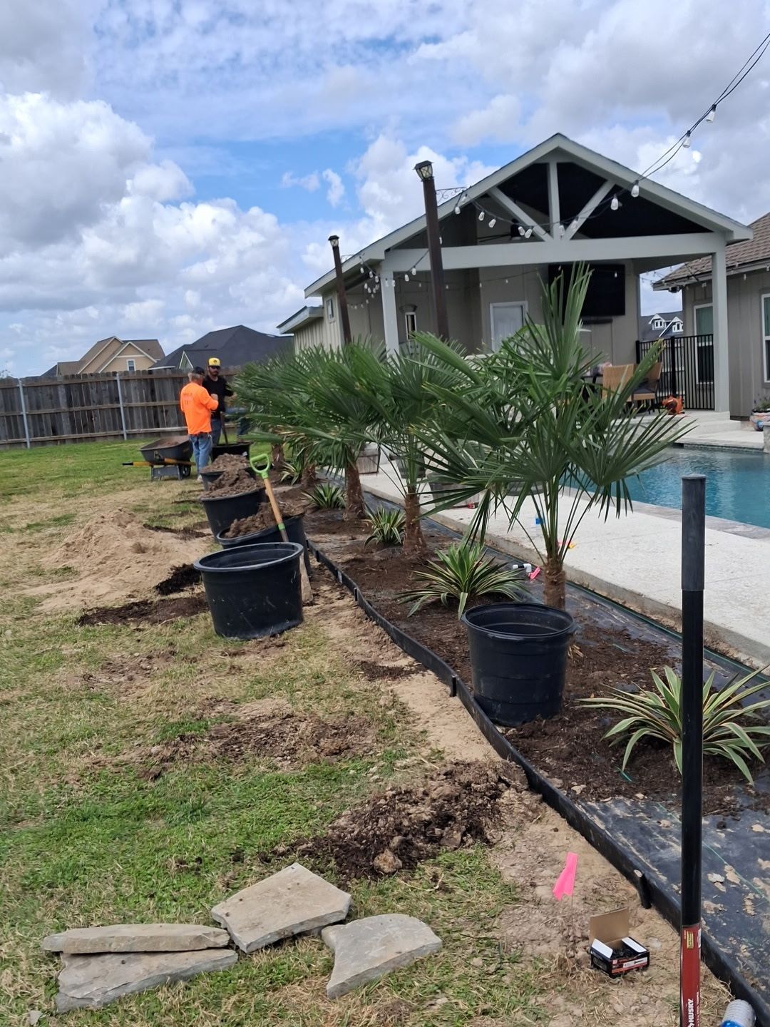 Landscaped yard with palm plants in pots, a worker in orange, and a house with a covered patio.