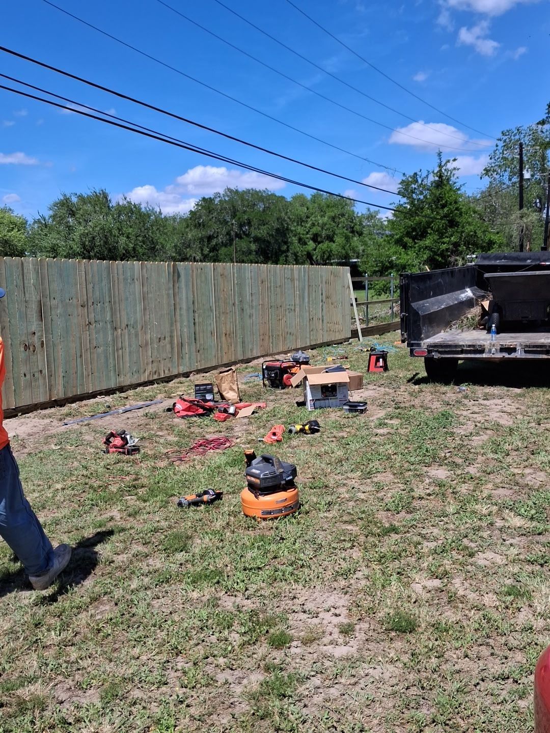 Backyard with scattered tools and debris near a wooden fence, car at right, and a person’s leg at left.