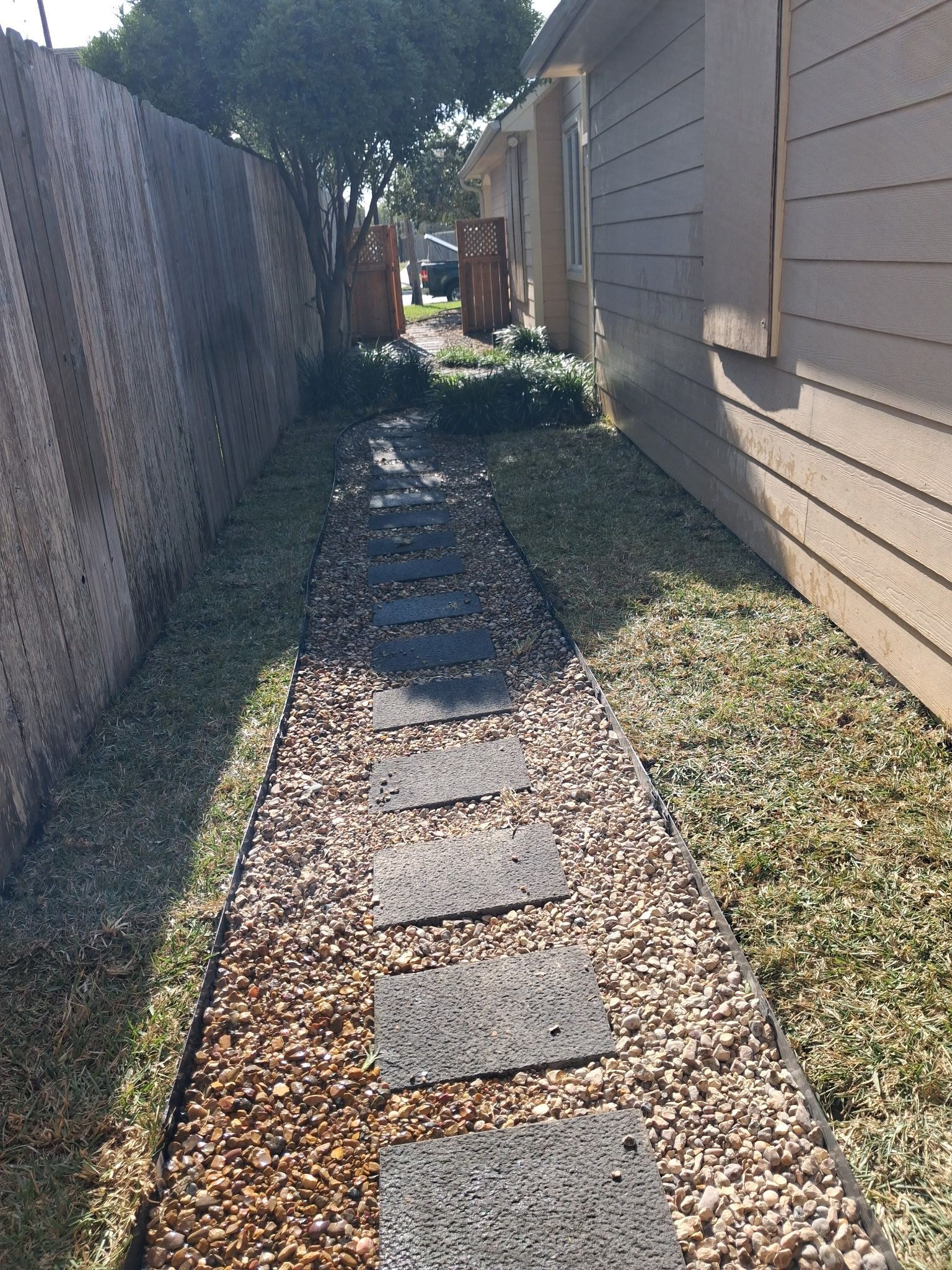 Narrow gravel path with stepping stones between a house and fence in sunlight