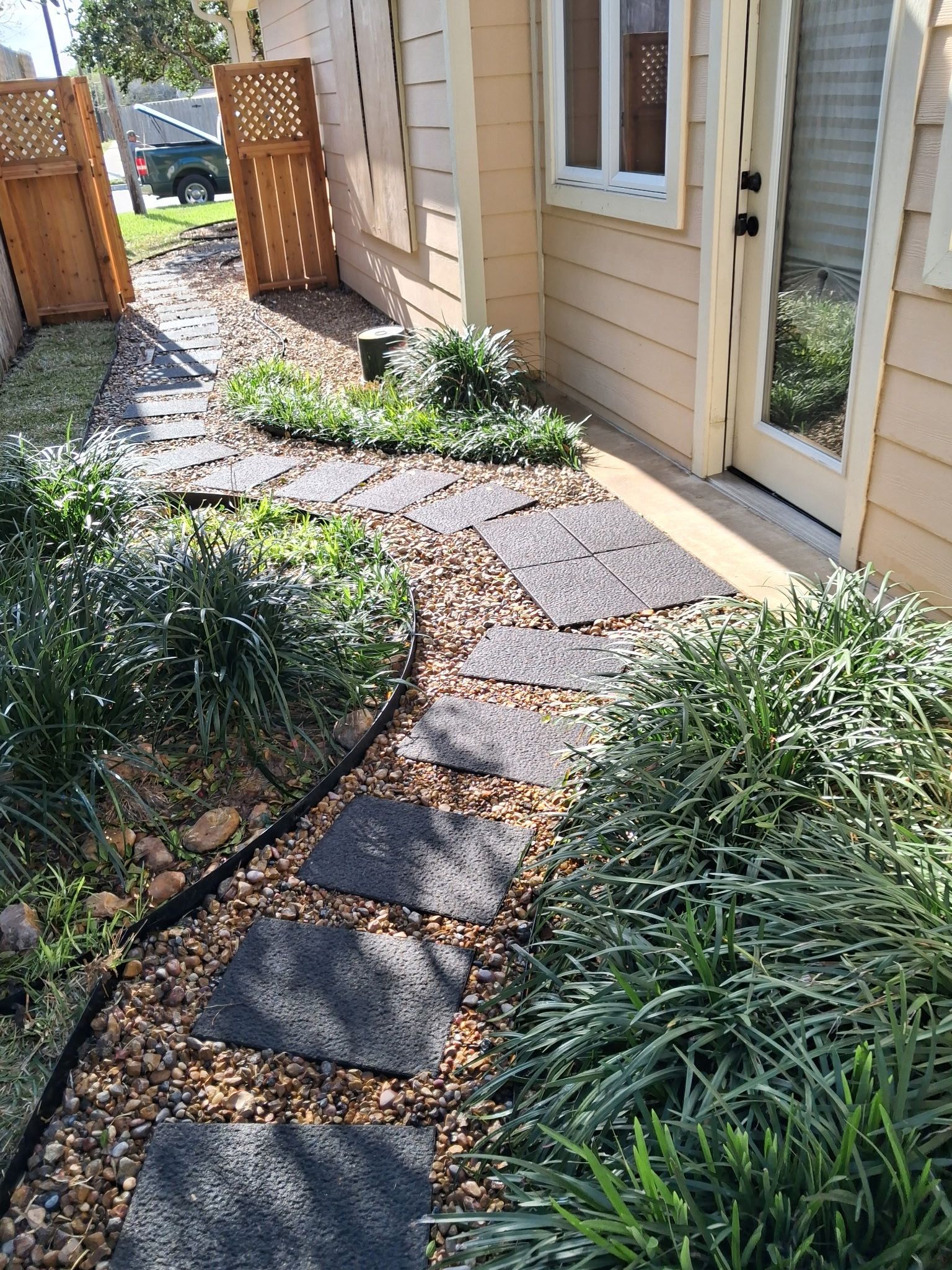 Narrow stone path beside a tan house, bordered by shrubs and mulch, leading to a wooden gate.