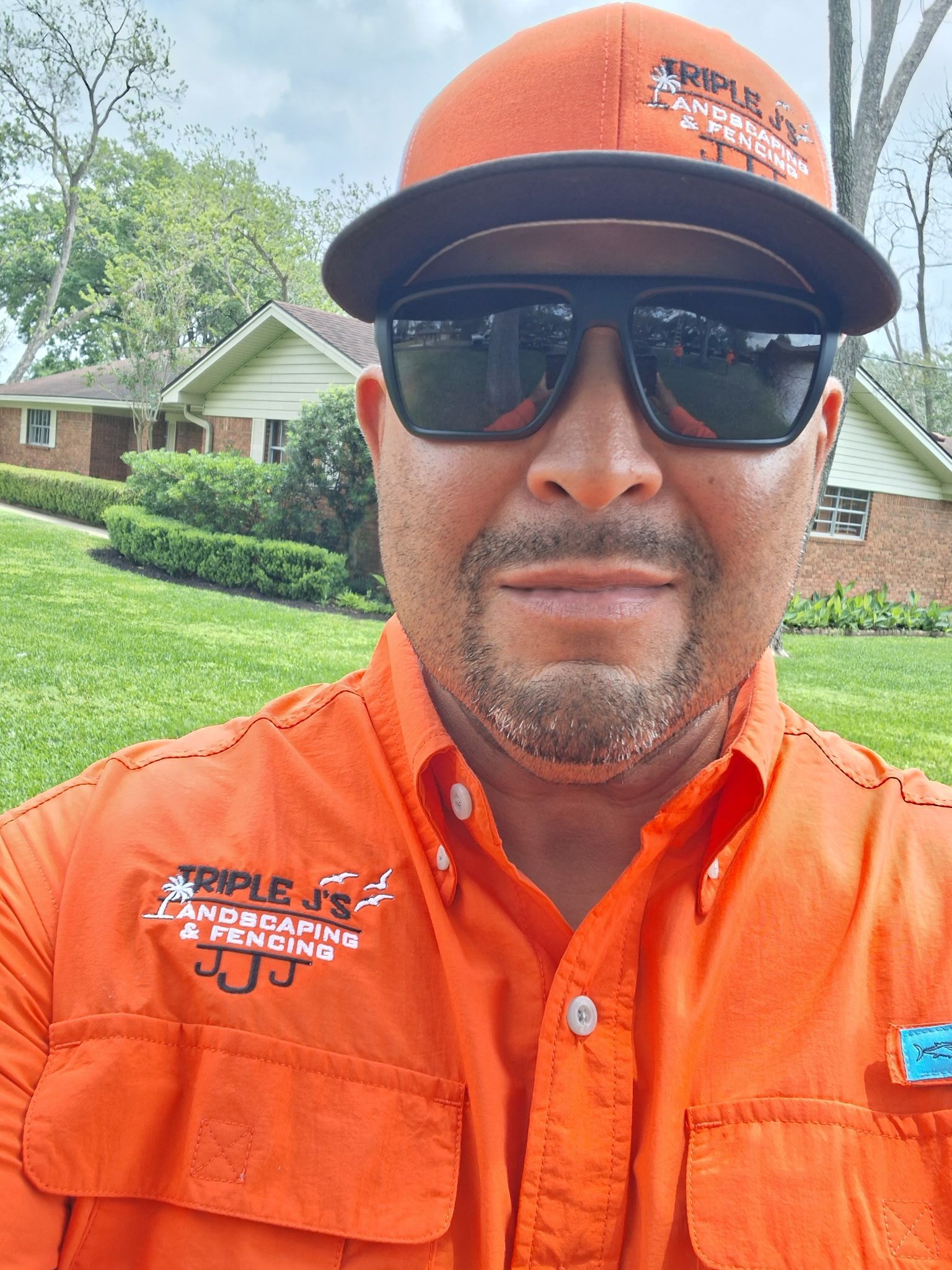 Man in orange work shirt, cap, and sunglasses taking a selfie in a suburban yard.