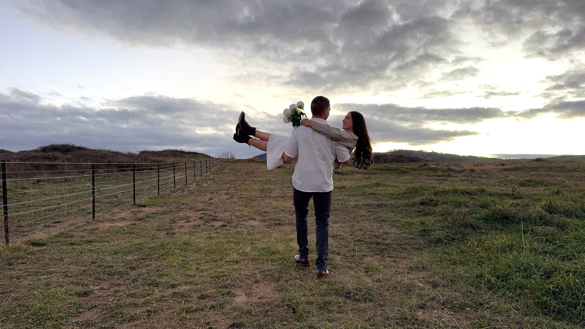 Nick carrying Chloe in a field at sunset, with a cloudy sky and fence line.