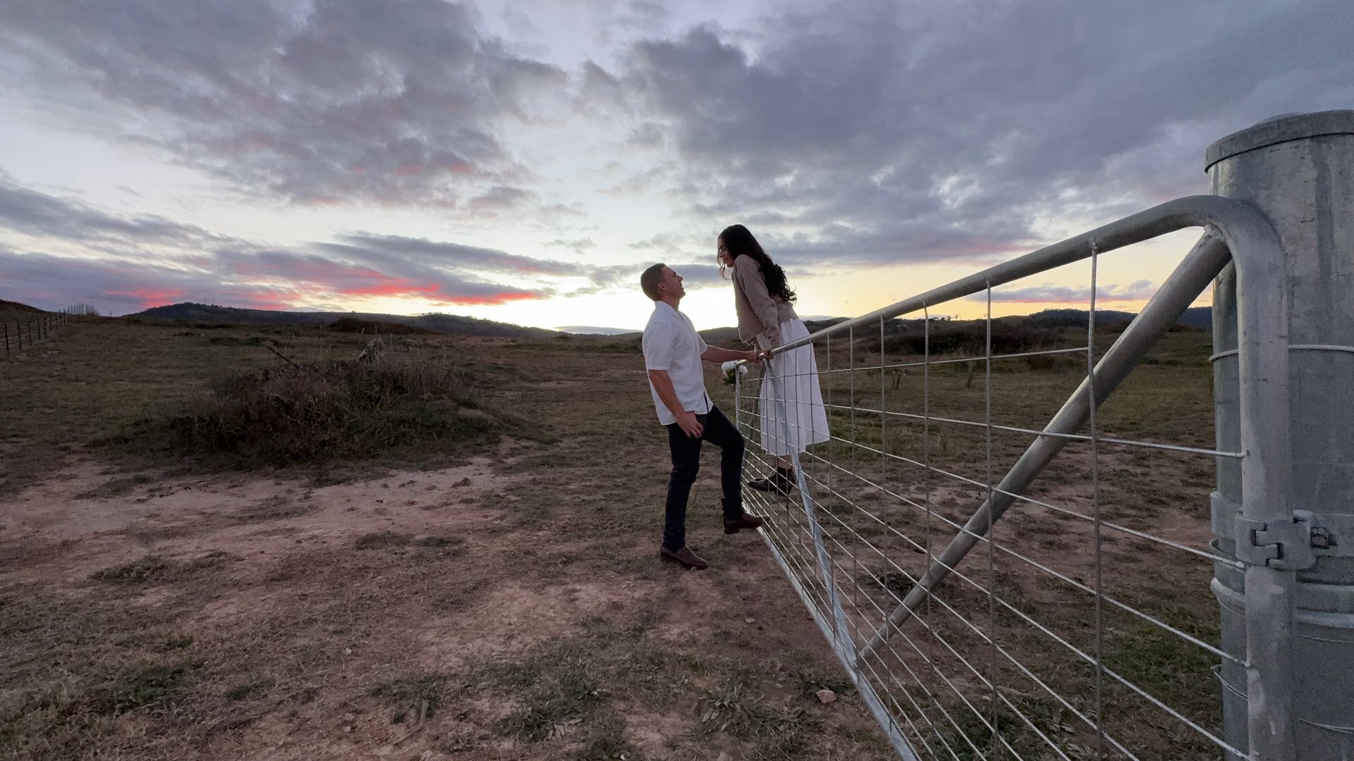 Nick and Chloe standing by a metal gate in a field at dusk under a cloudy sky