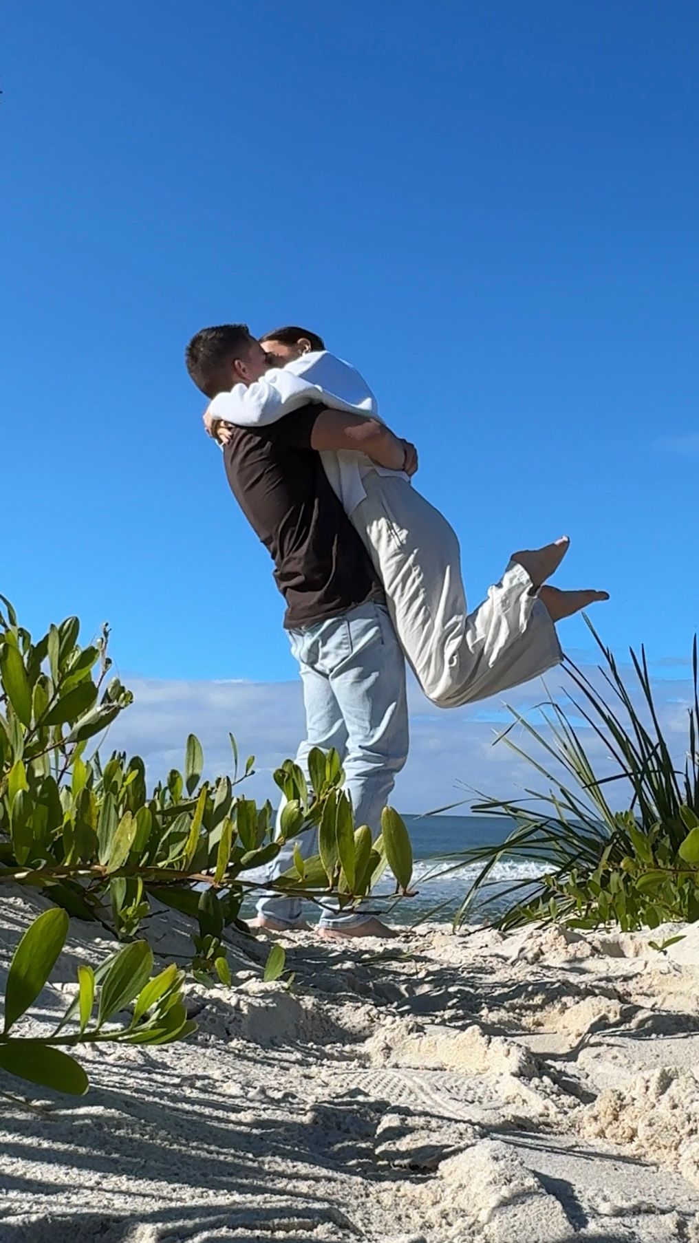 Nick and Chloe people hugging and jumping on a sunny beach under a clear blue sky