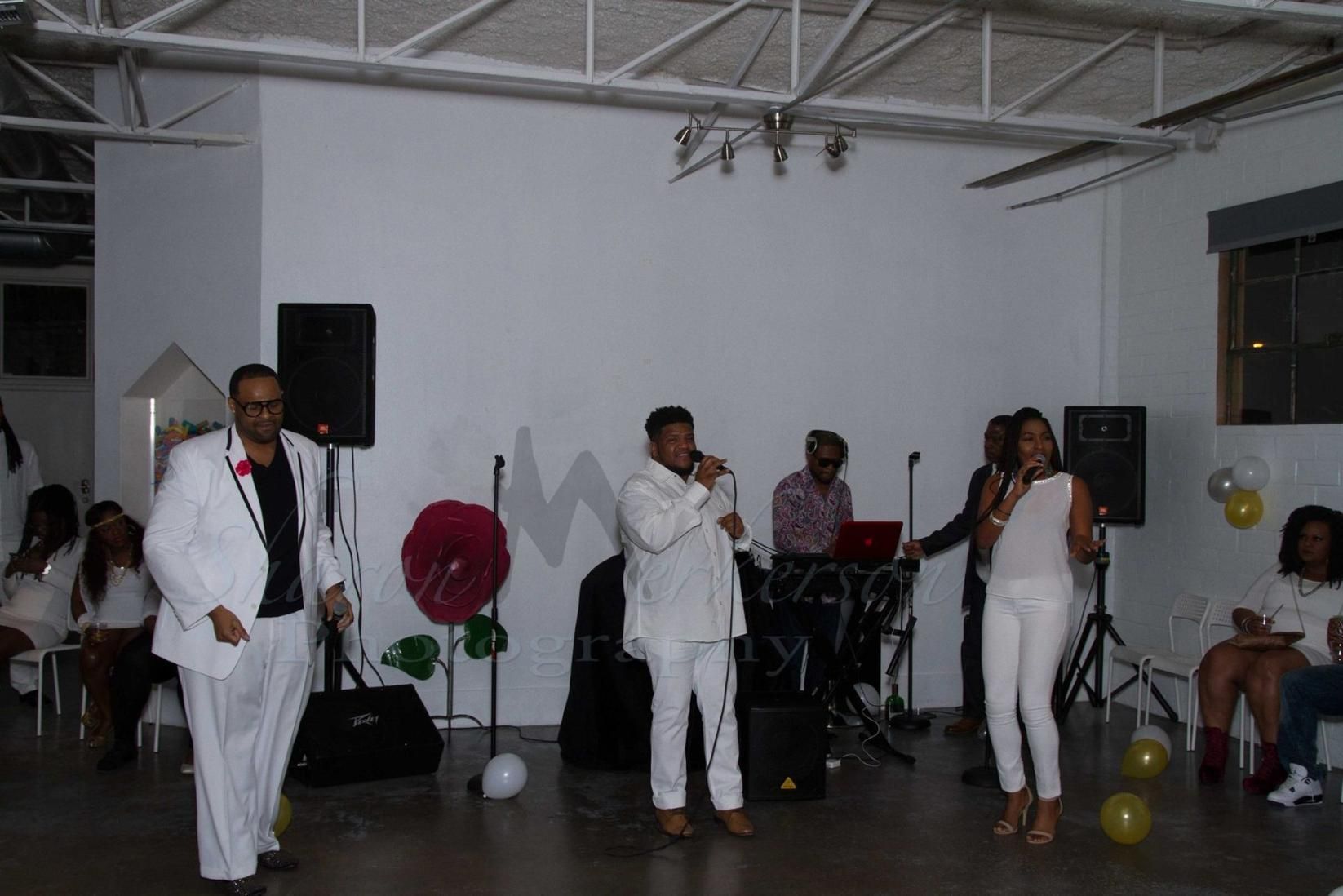 People on a small stage performing with microphones and instruments in a white indoor venue