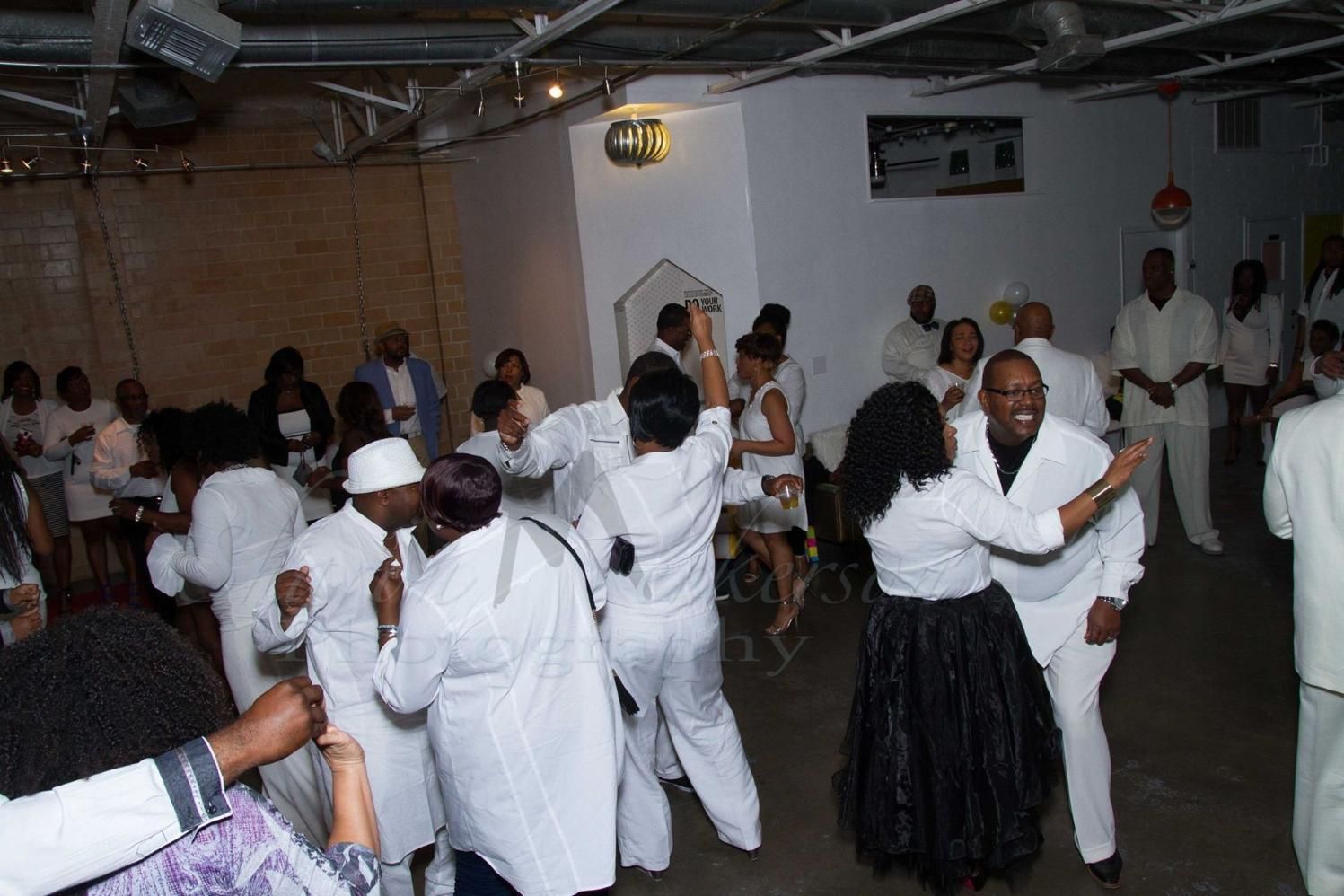 People dressed in white dancing at an indoor celebration with a crowd watching nearby