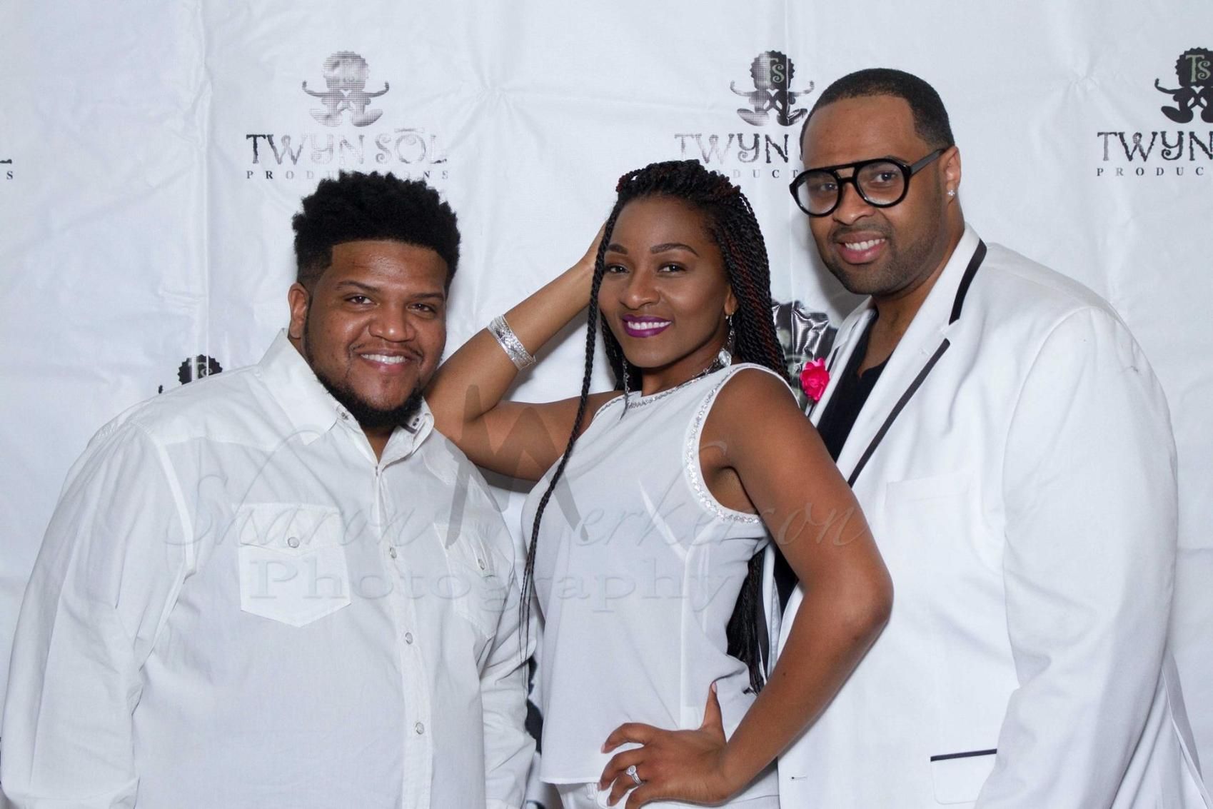 Three people posing in white outfits at a step-and-repeat event backdrop
