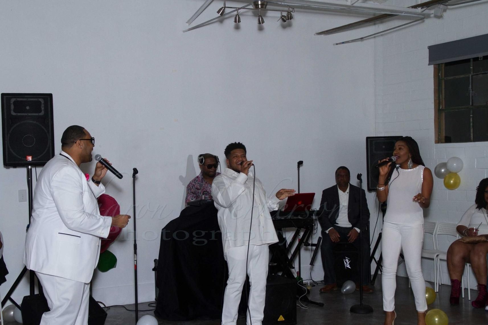 Three singers in white perform onstage with a small band in the background