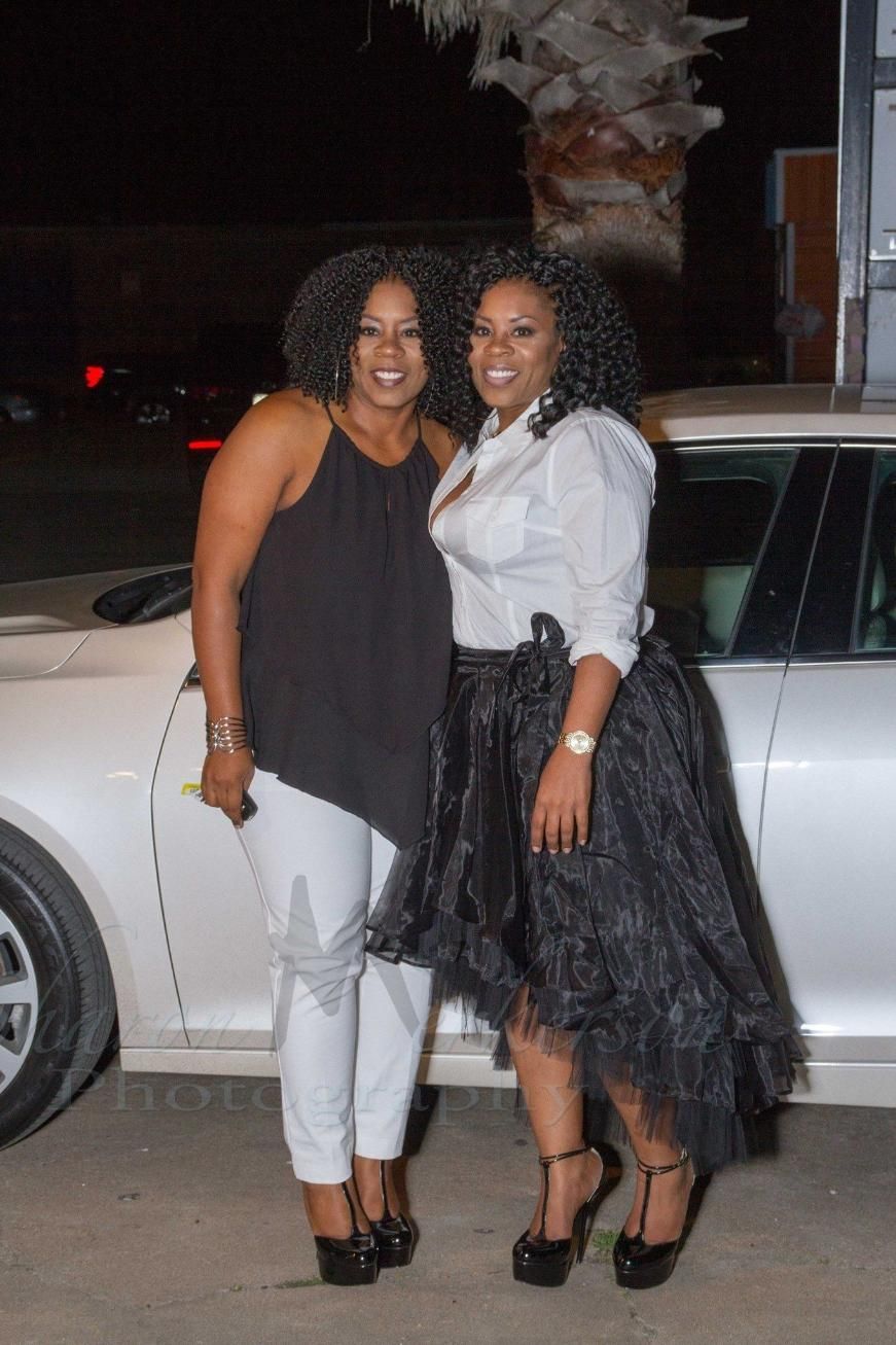 Two women posing outdoors at night beside a silver car, both smiling and dressed in black-and-white outfits
