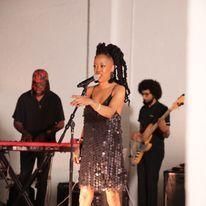 Singer in a sparkly dress performing onstage with a keyboardist and bassist behind her.