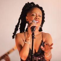 Singer performing into a microphone on stage, wearing a black dress with long braided hair.