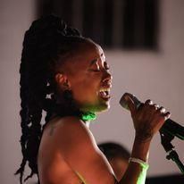 Singer performing into a microphone on stage, wearing a green choker and bracelet, eyes closed.