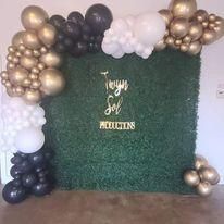 Gold and black balloon arch around a green hedge backdrop with gold lettering at an event backdrop.