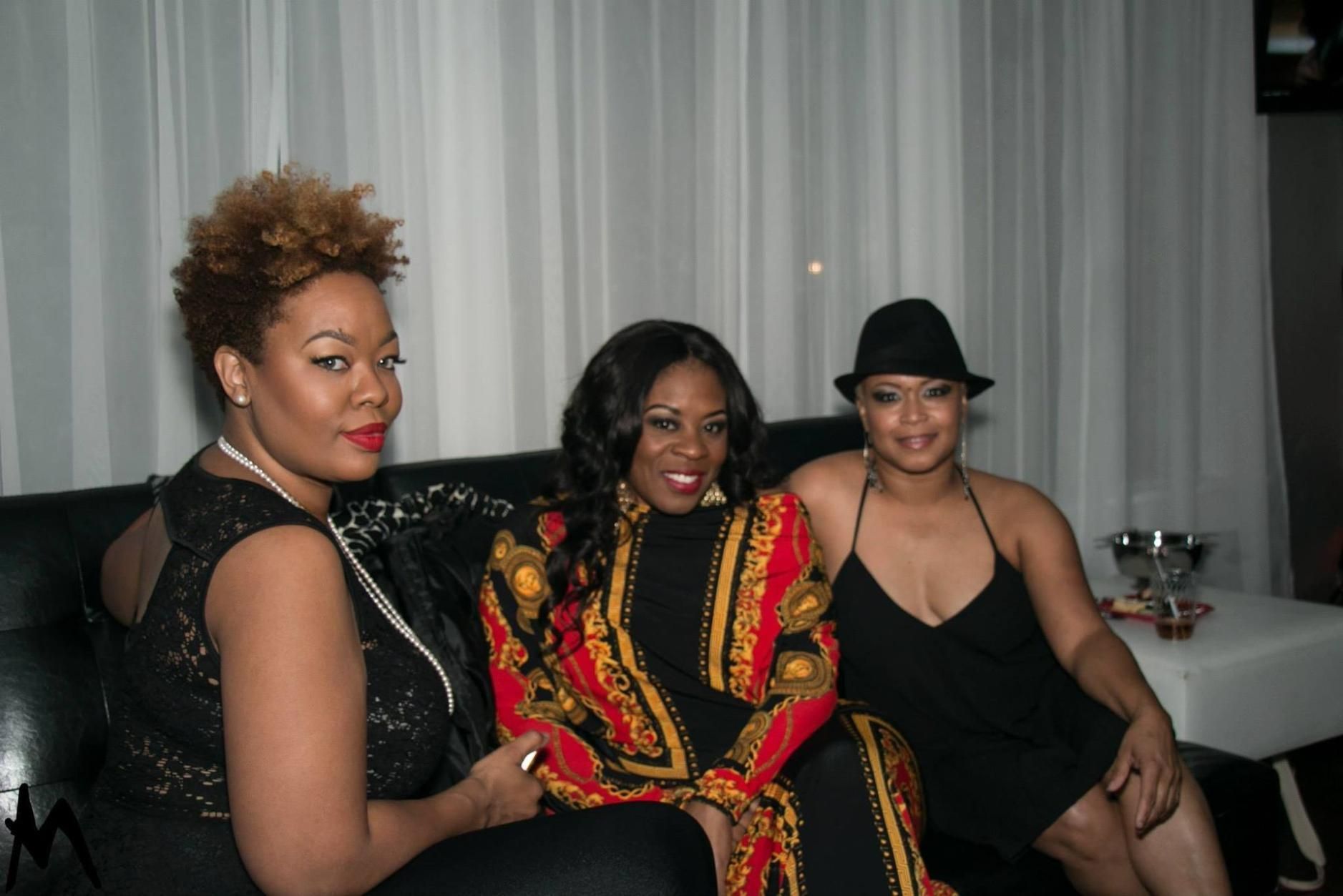 Three people seated on a couch, posing at an indoor event with white curtains and a table nearby