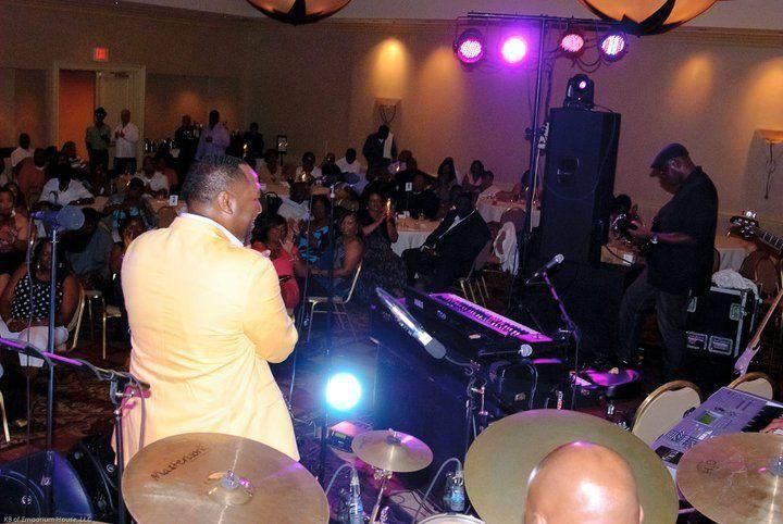 Live band performance on a stage with a singer in a yellow jacket facing a seated audience under purple lights