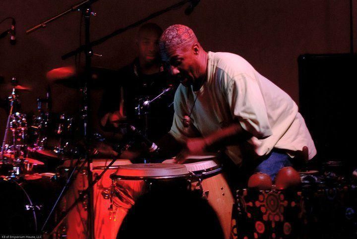 Musician playing hand drums onstage under red lights, with a drummer blurred in the background.